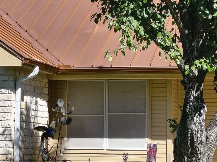 Brown gutter below a copper-colored metal roof with a white downspout beside the window